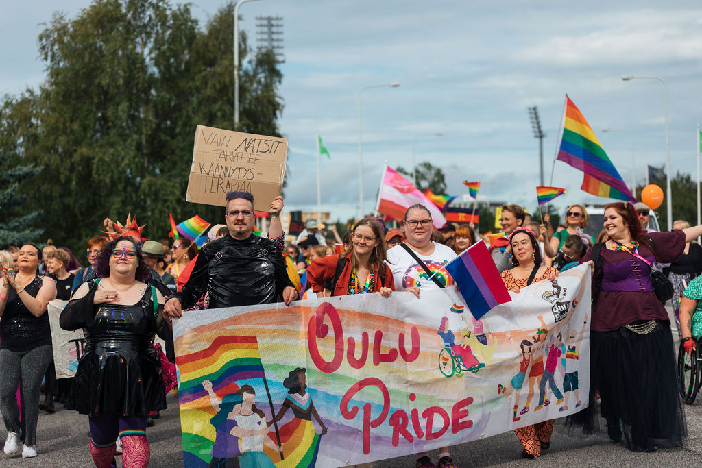 Ihmisiä marssimassa iloisena kulkueessa. Kuvassa etualalla kävelevillä on banneri, jossa lukee Oulu Pride