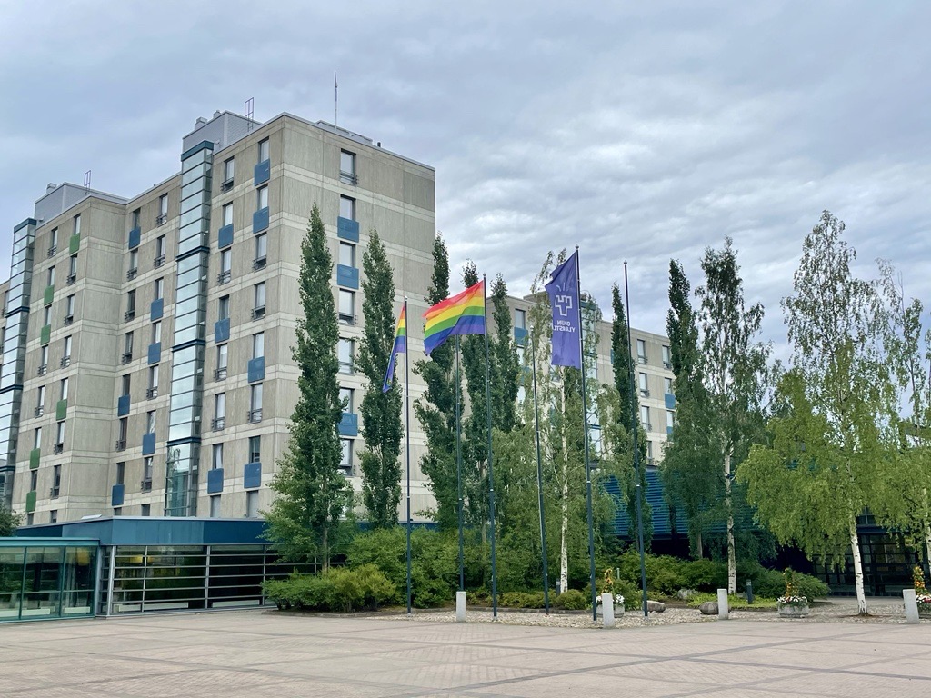 Edustalla lipputankoja, joissa Oulun yliopiston lippu ja kaksi sateenkaarilippua. Taustalla Oulun yliopiston rakennuksia.